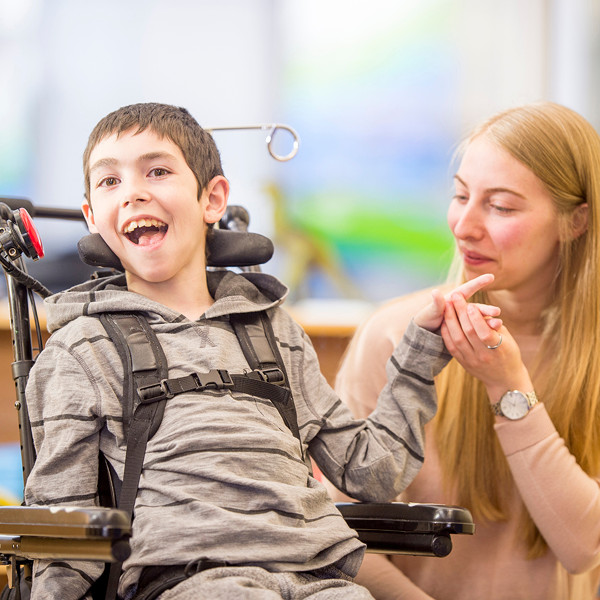 Child in a supportive wheelchair holding hands with a caregiver during a therapy session.