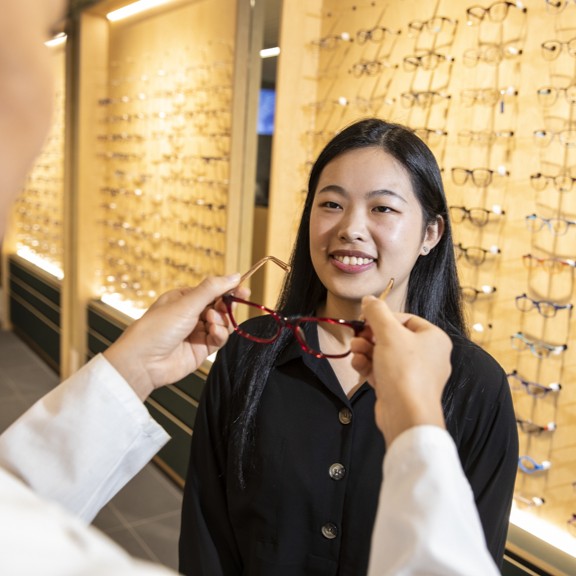 young woman with dark hair and black button up shirt standing in front of optometry clinic spectacles showcase wall, receiving a pair of spectacles from a clinician in a white clinical gown